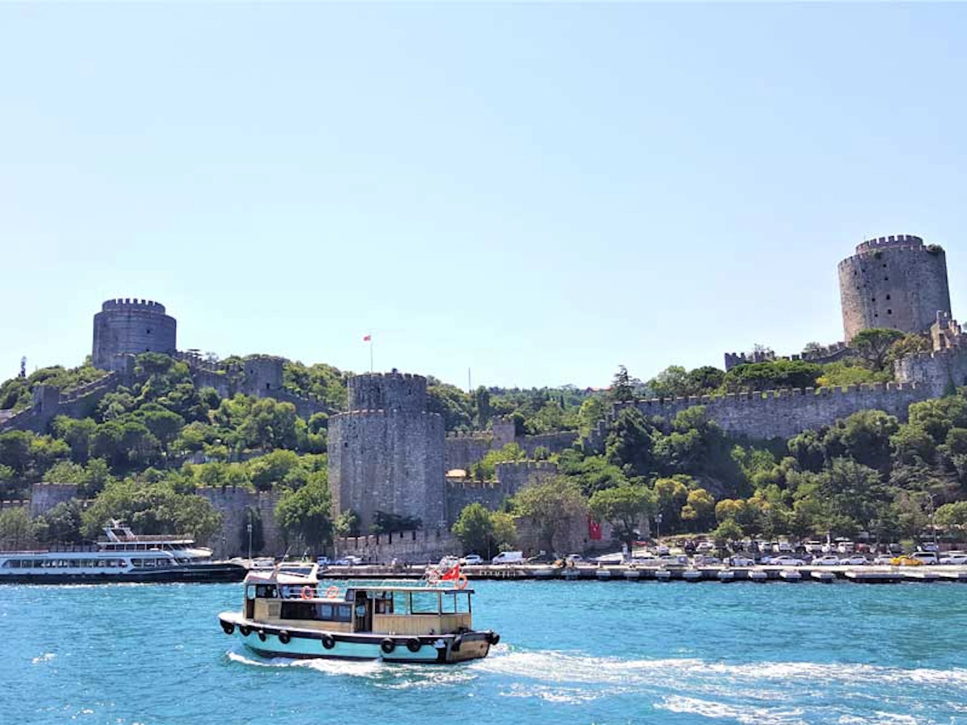 A small boat sails on a blue river in front of a historic fortress on a green hillside under a clear sky.