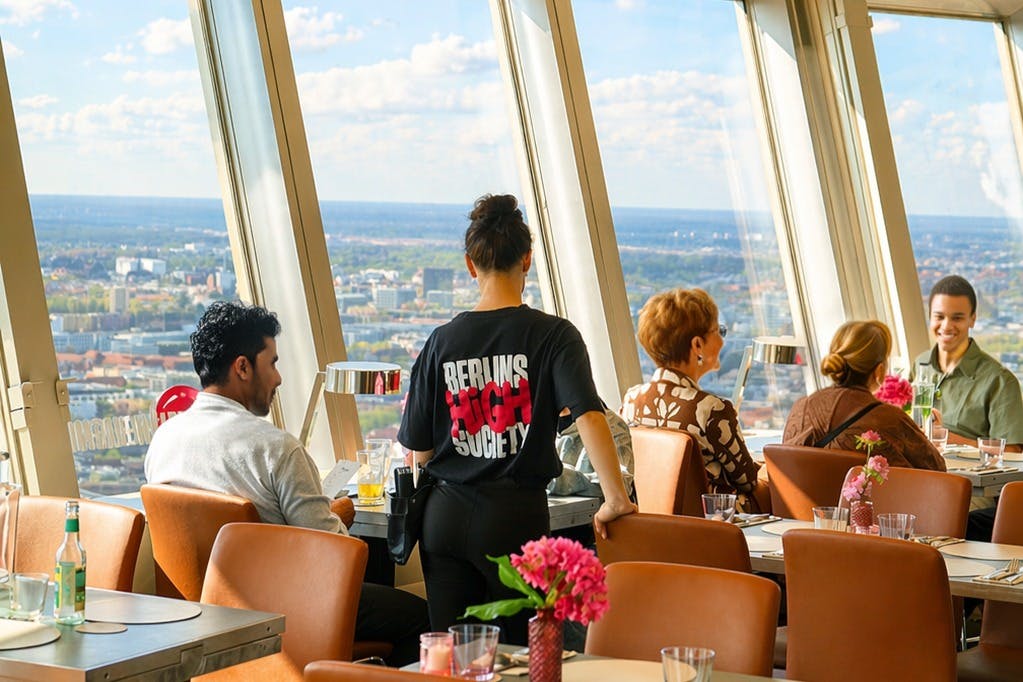 Ein Restaurant mit großen Fenstern, durch die man einen Blick auf die Stadt hat. Die Gäste sitzen an Tischen, und ein Kellner geht vorbei.
