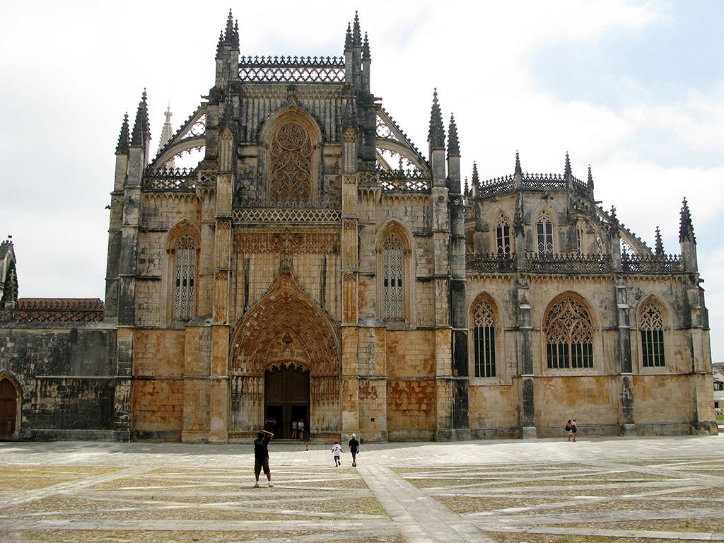 曇りの日には、尖ったアーチと細かい石の彫刻が施された、大きくて華やかなゴシック様式の大聖堂に向かって歩く人々がいます。