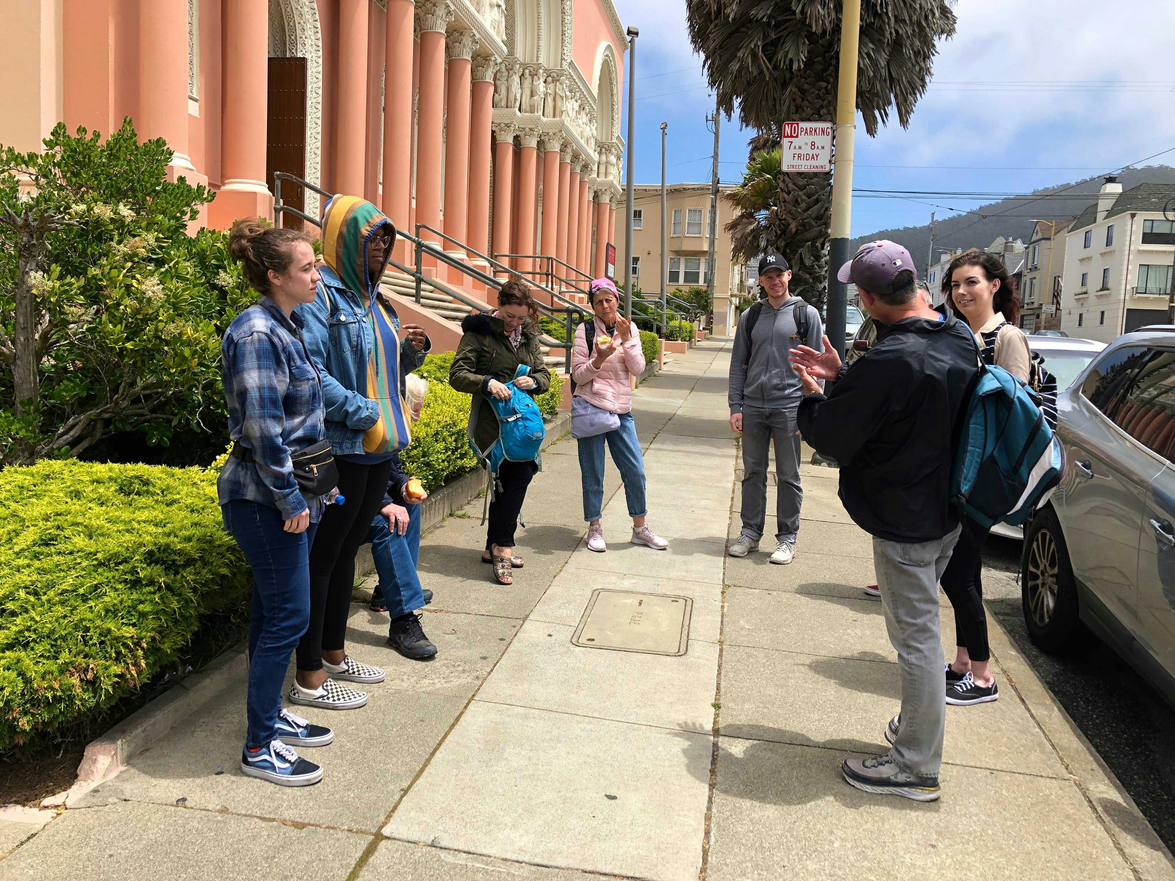 A tour guide speaks to a group of people standing on a sidewalk near a pink building with arches.