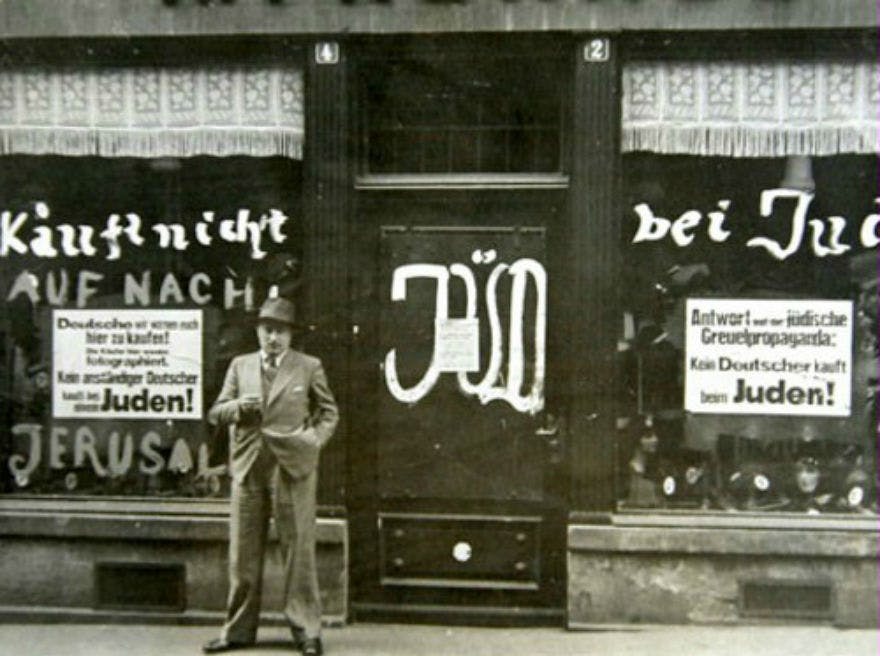 Ένας άνδρας με κοστούμι στέκεται έξω από ένα κατάστημα με τα παράθυρα και την πόρτα του βανδαλισμένα με αντισημιτικά γκράφιτι στα γερμανικά.