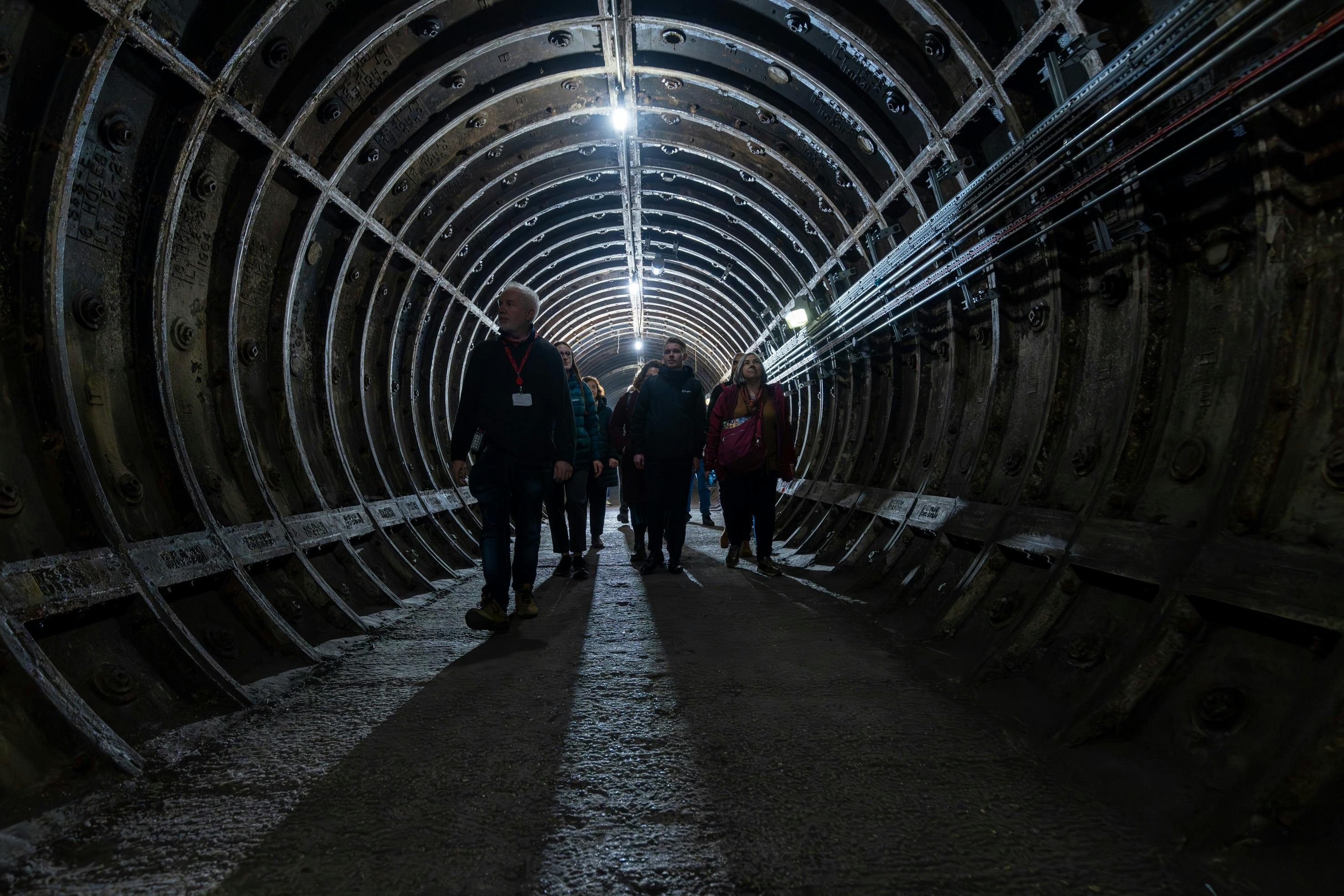Eine Gruppe von Menschen geht durch einen schwach beleuchteten unterirdischen Tunnel mit gebogenen Metallwänden und Oberlichtern.