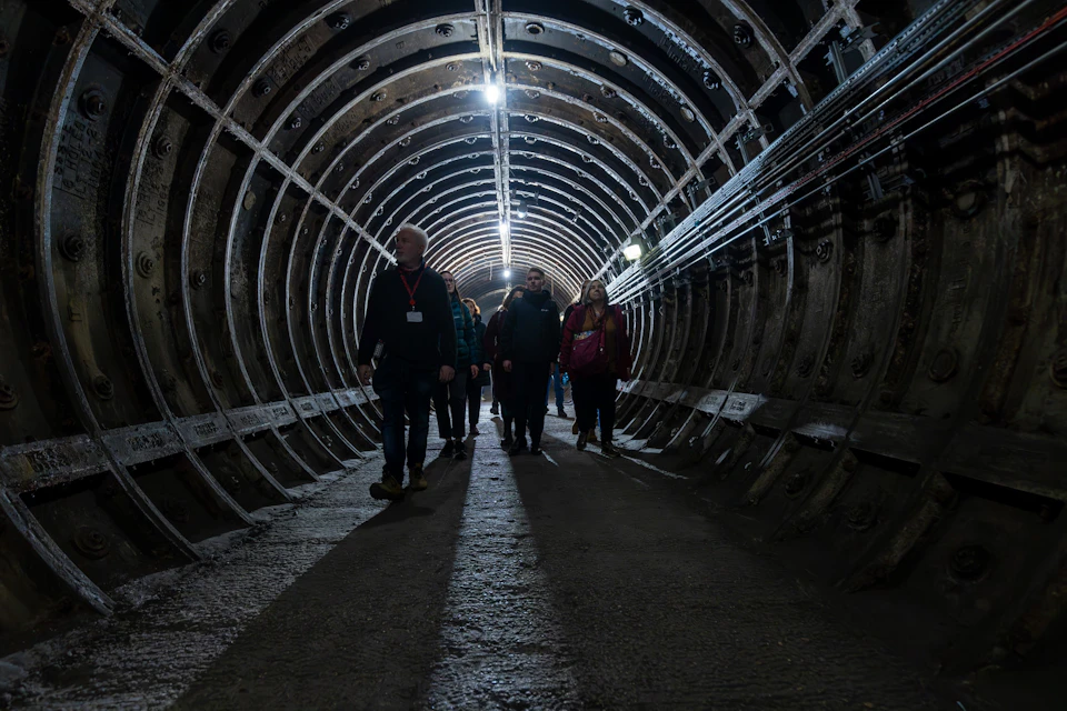 A group of people walking through a dimly lit underground tunnel with curved metal walls and overhead lights.