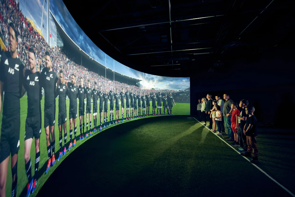 A group of people viewing a large curved screen displaying a rugby team lined up on a field during a match.
