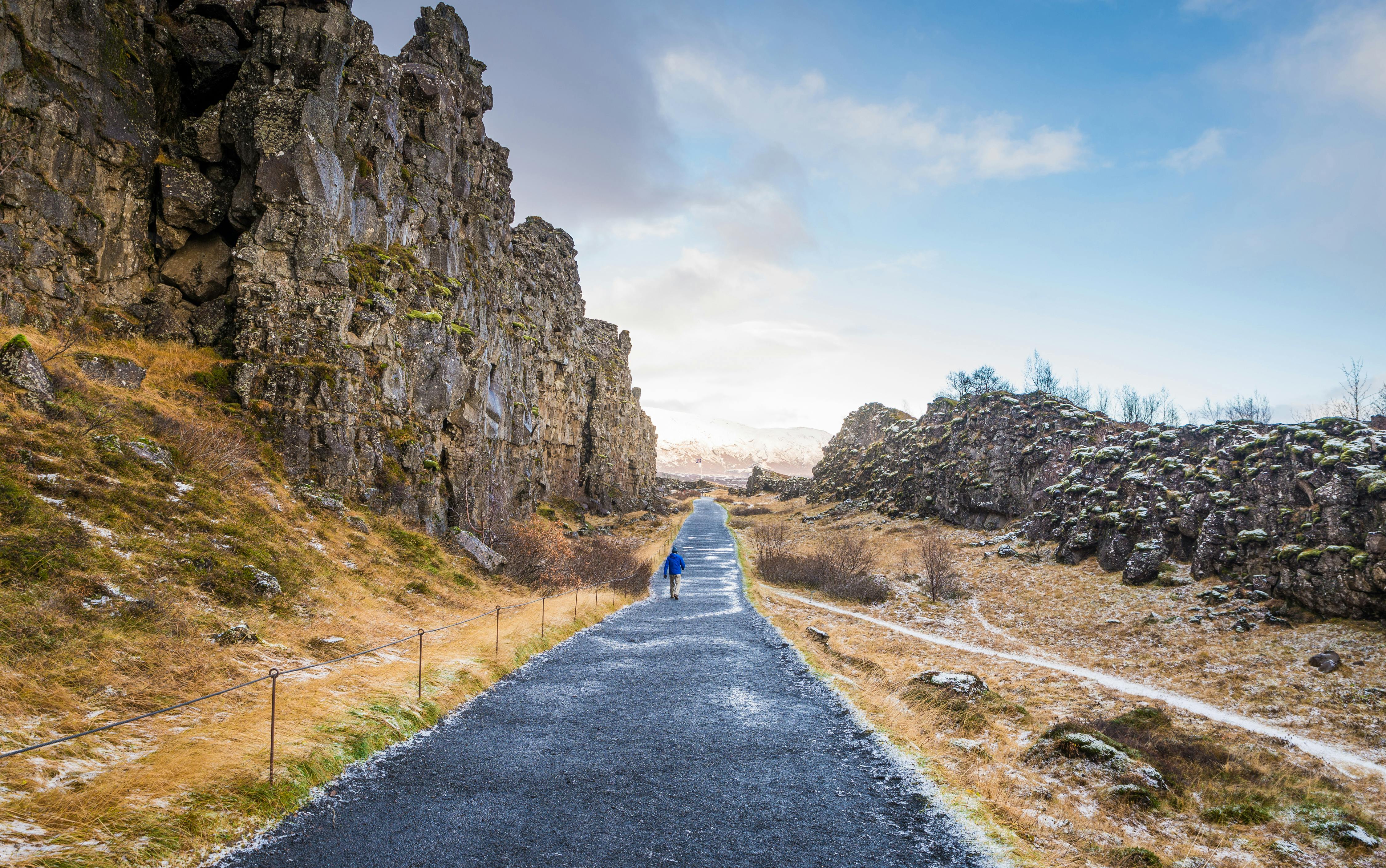 Člověk kráčí po dlážděné cestě lemované skalnatými útesy a suchou trávou pod částečně zataženou oblohou.
