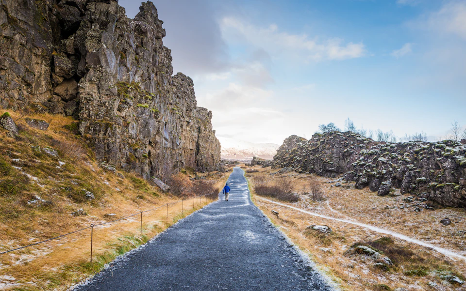 A person walks along a paved path flanked by rocky cliffs and dry grass under a partly cloudy sky.