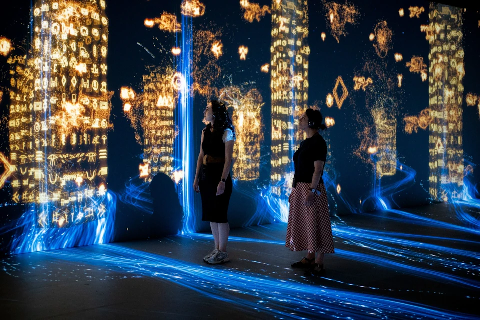 Two people standing in a dark room with illuminated abstract designs and symbols projected on the walls and floor.