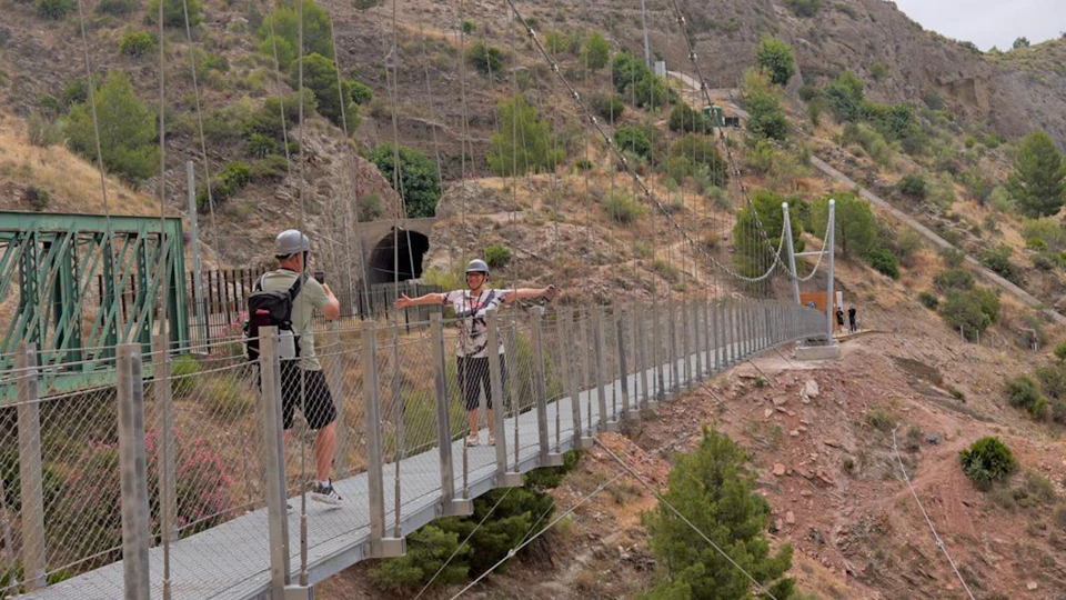 Two people wearing helmets on a suspension bridge in a rocky and green landscape, with one person posing for a photo.