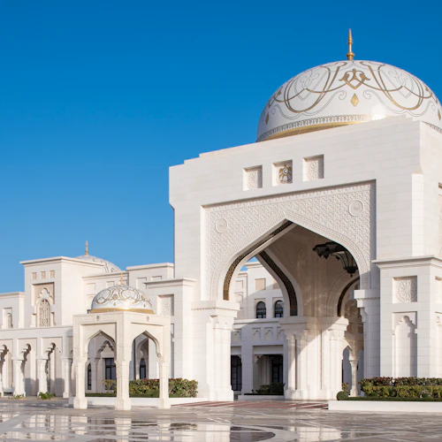 Weißes Gebäude mit verschnörkelten Kuppeln, Bögen und dekorativen Details vor einem klaren blauen Himmel. Innenhof mit reflektierendem Bodenbelag.