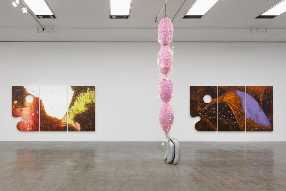 A gallery space with abstract artworks on the walls and a tall hanging sculpture made of pink and metallic elements in the center.