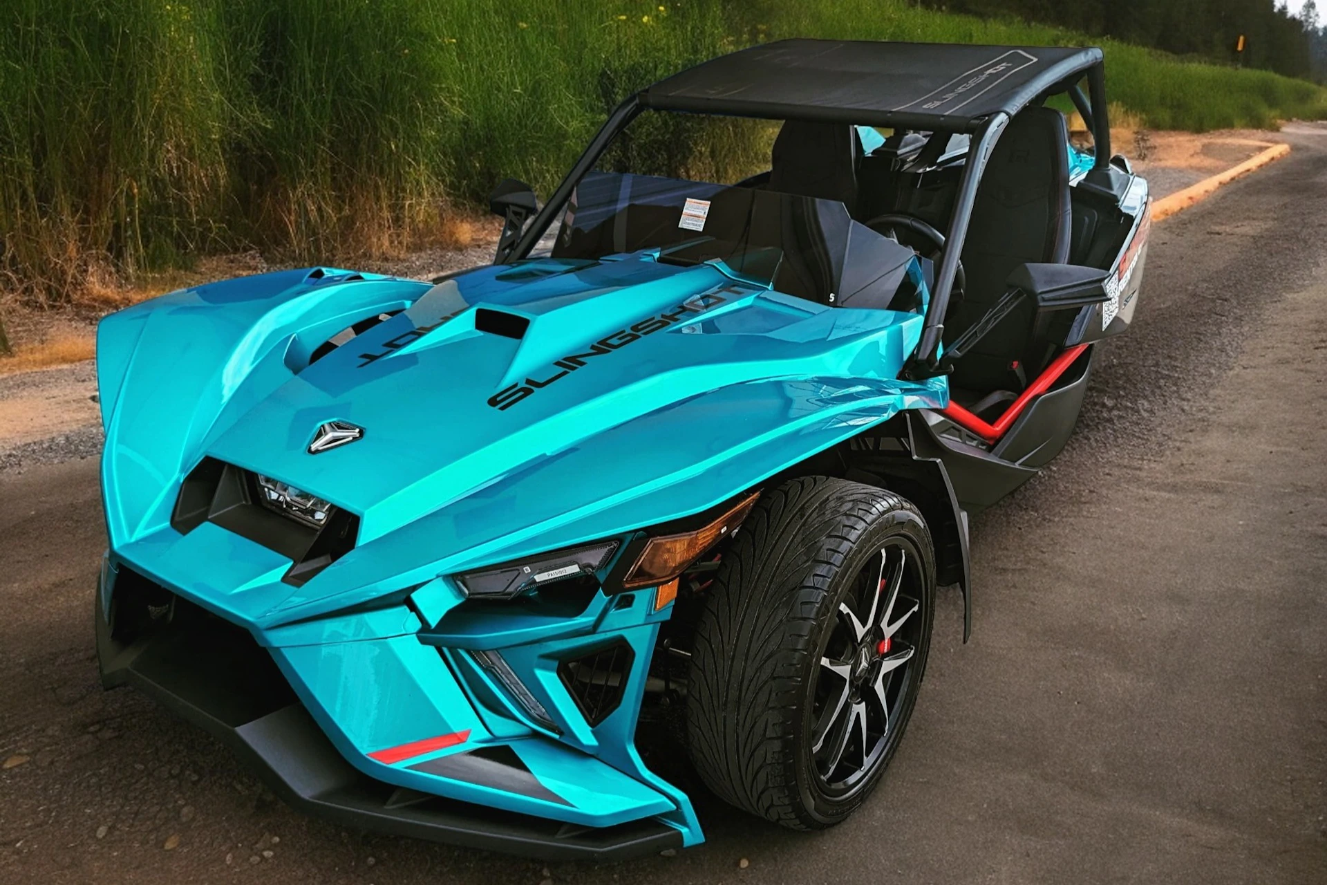 A bright blue three-wheeled Polaris Slingshot vehicle parked on a road with grassy surroundings.