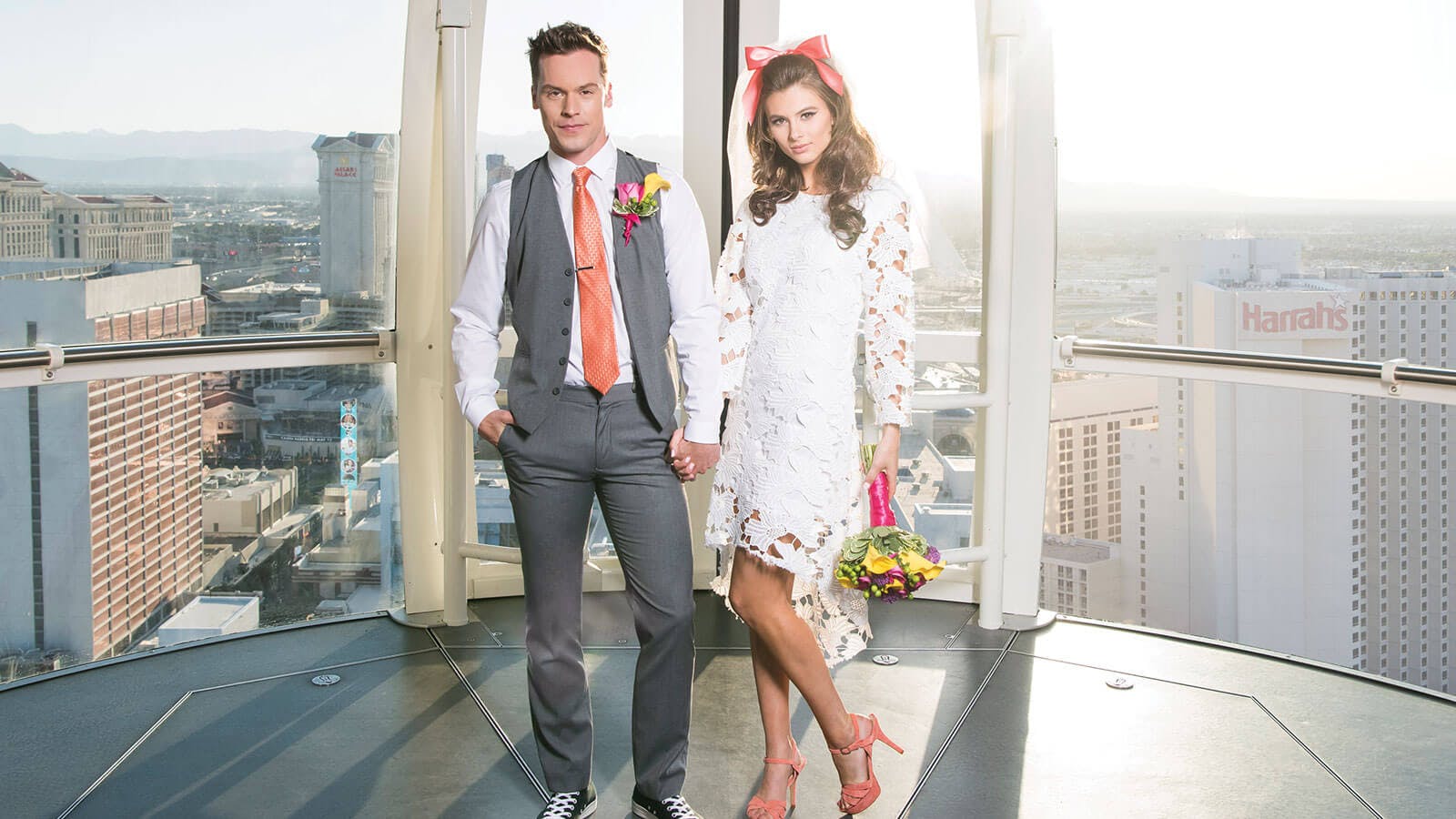 A couple in wedding attire stands in a high-rise with a cityscape view; the man in a gray suit and orange tie, the woman in a white dress.