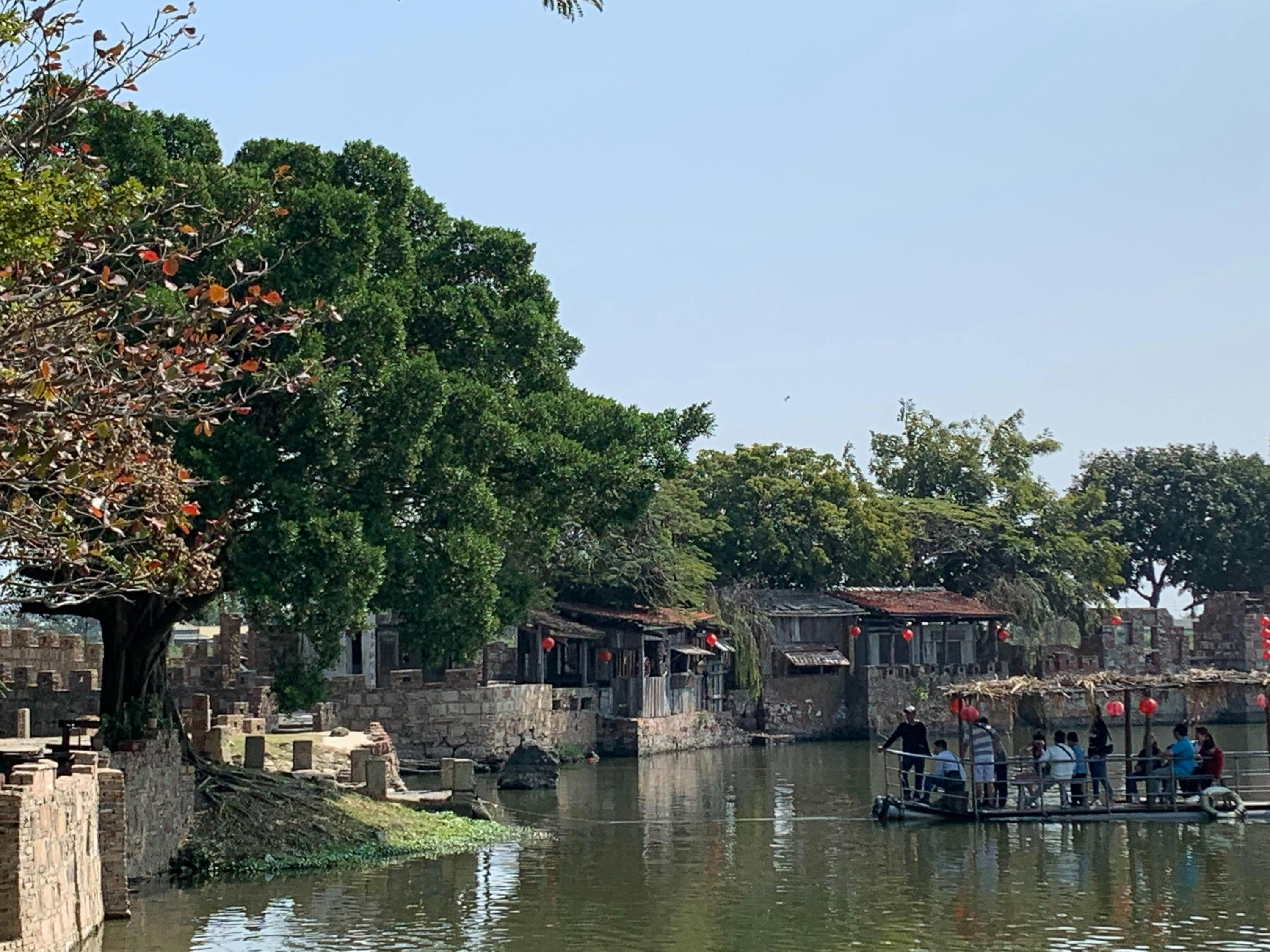 A serene riverside scene with rustic houses, trees, and people on a small boat under blue skies.
