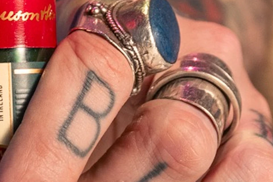 A close-up of a Jameson whiskey bottle on a table, with a tattooed hand adorned with rings pointing at it.