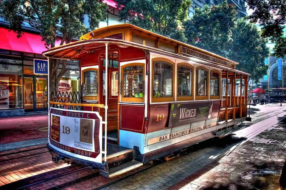 A red and yellow San Francisco cable car with the number 19, next to trees and buildings, promoting the musical Wicked.