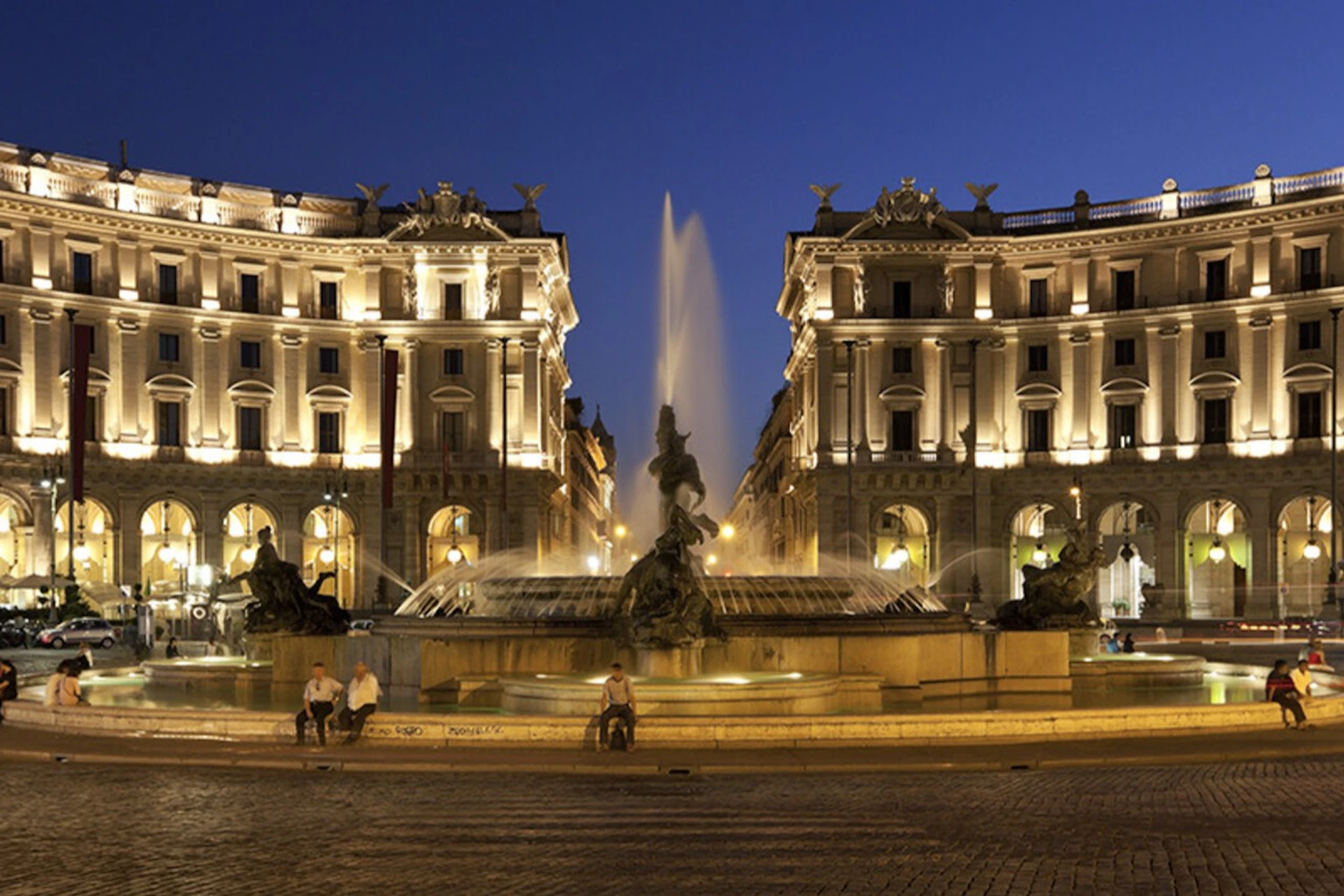 Illuminated buildings and a fountain with statues at night, with a few people seated nearby in a public square.