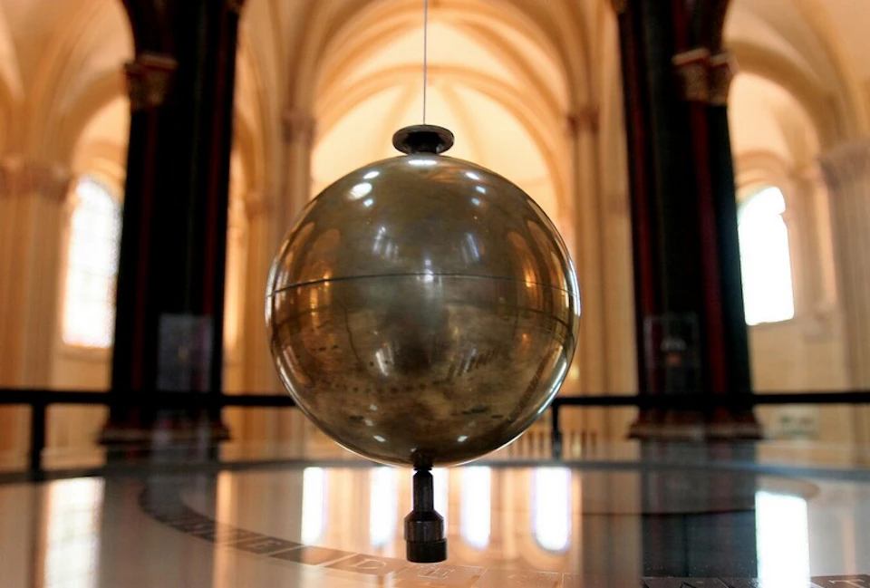 A large, shiny pendulum hanging inside a grand building with arched ceilings and ornate columns in the background.