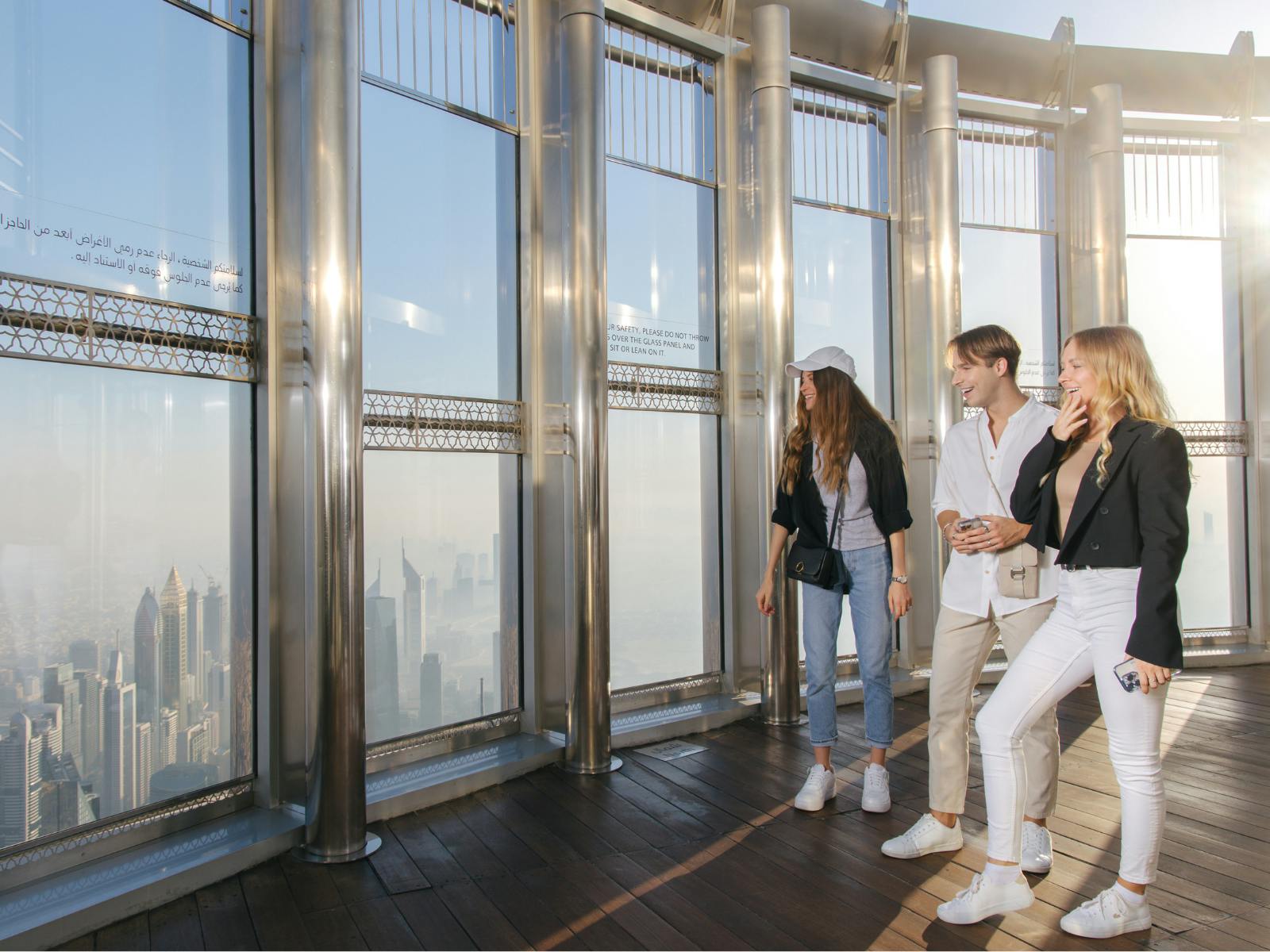 Three people stand on an observation deck with large windows, smiling and enjoying the view of a city skyline in the background.