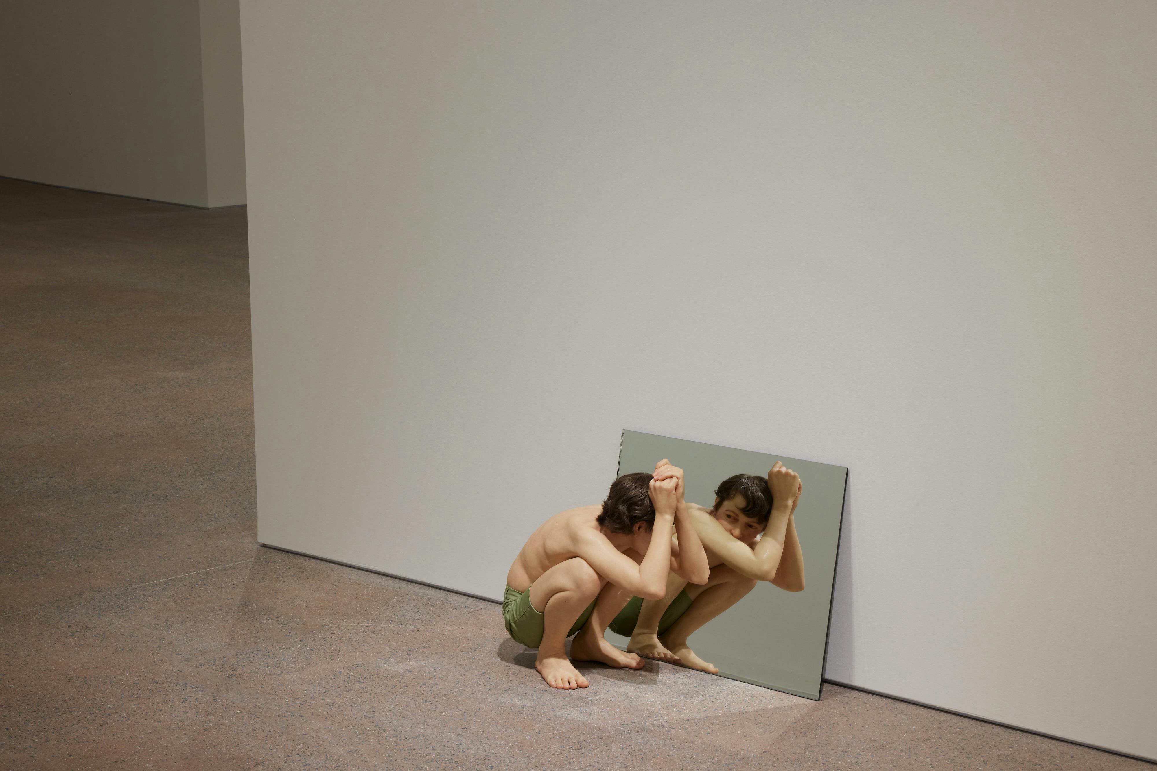 A figure crouching near a mirror on the floor, reflecting their image. The setting is a minimal interior space.