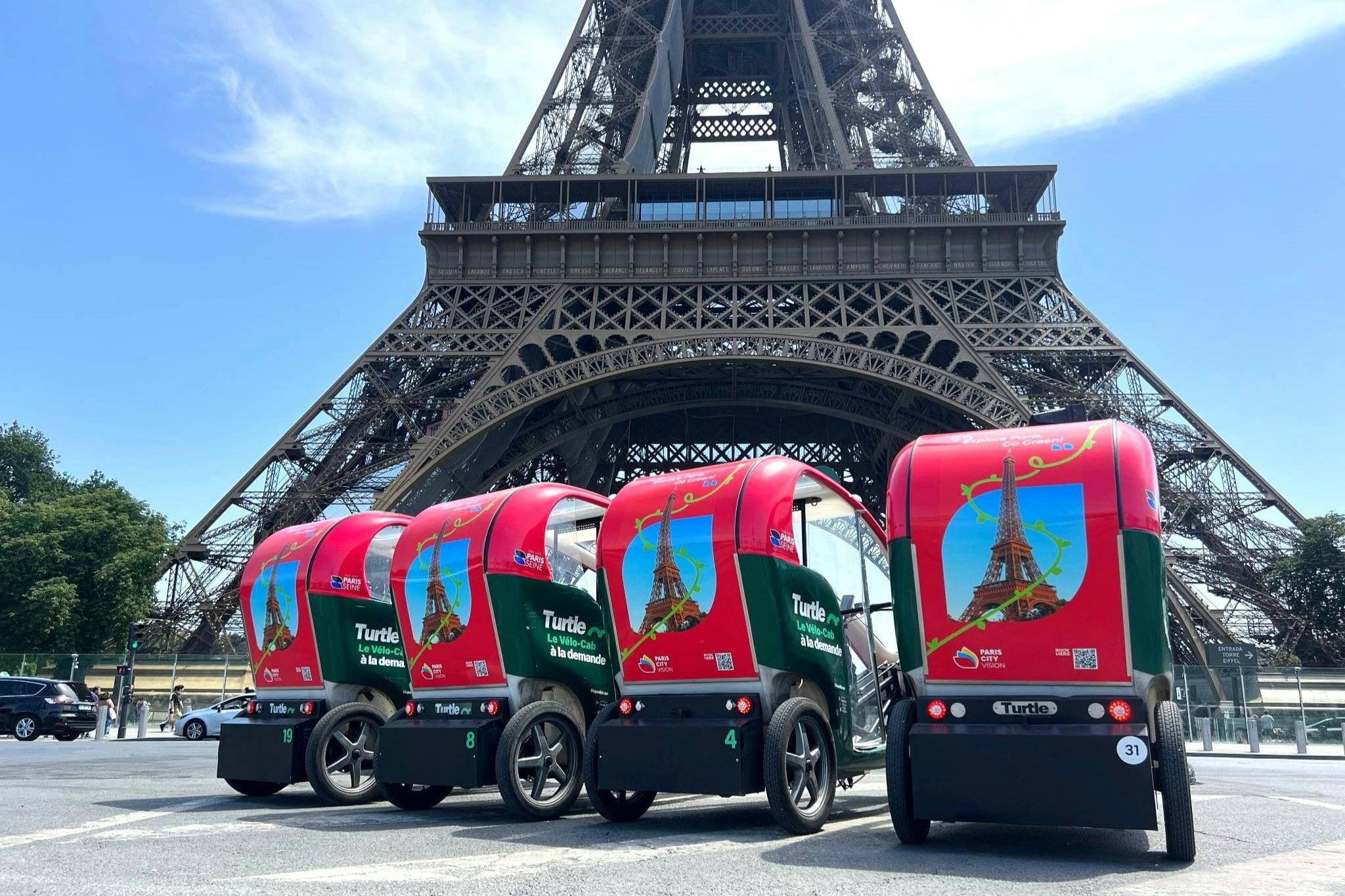 Четыре небольших красных и зеленых электромобиля припаркованы бок о бок перед Эйфелевой башней в ясный день.