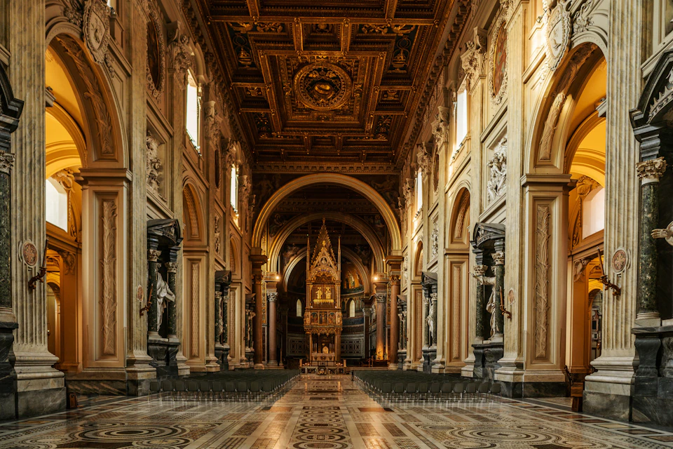 An ornate church interior with a patterned floor, arched ceiling, statues, and a decorated altar in the background.
