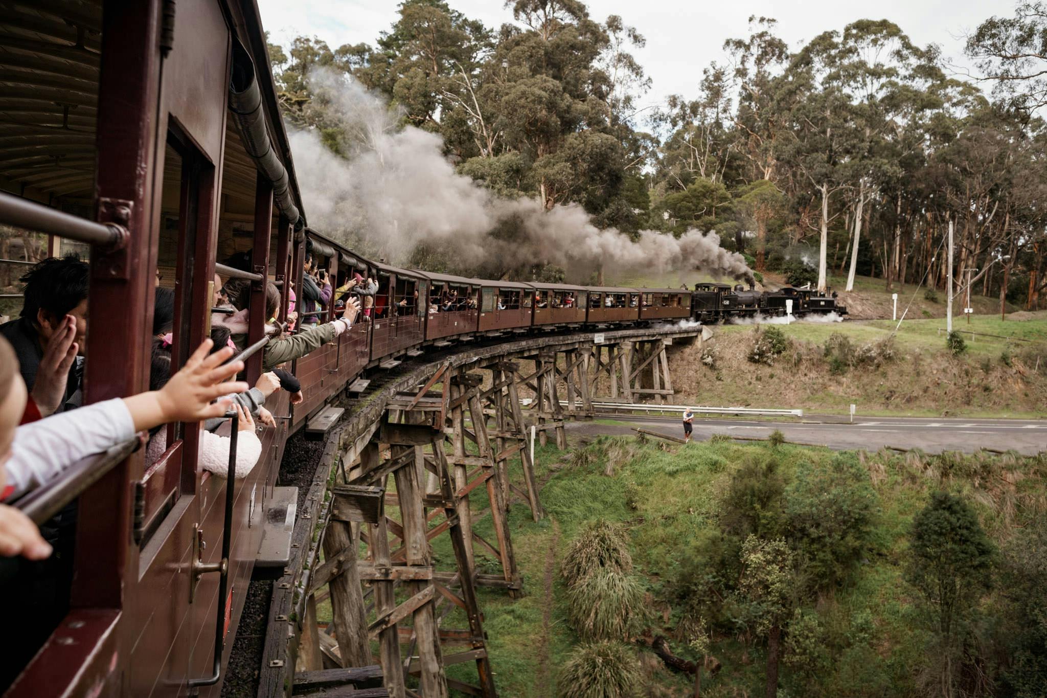 Un train à vapeur traverse un pont à chevalets en bois, les passagers font des signes de la main, entourés d'arbres et de verdure.