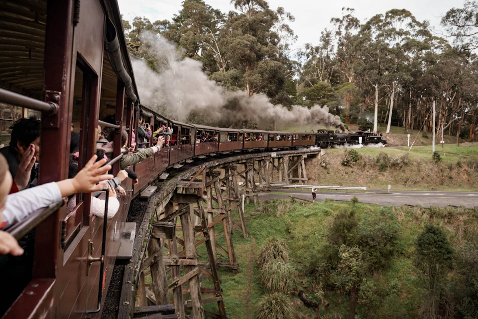 A steam train crossing a wooden trestle bridge, passengers waving, surrounded by trees and greenery.