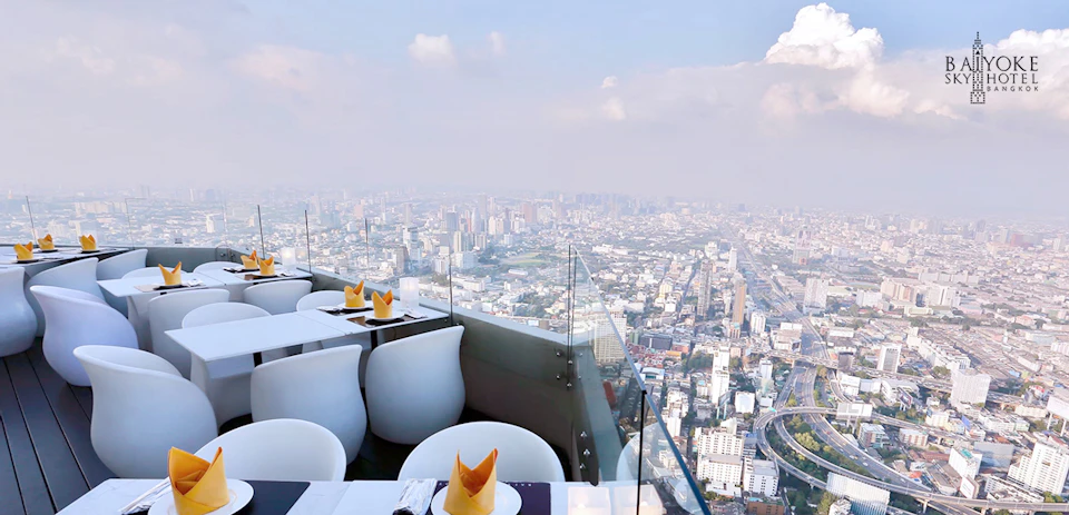 Outdoor rooftop restaurant with white chairs, tables set with napkins, and a glass barrier, offering a panoramic view of a city skyline.