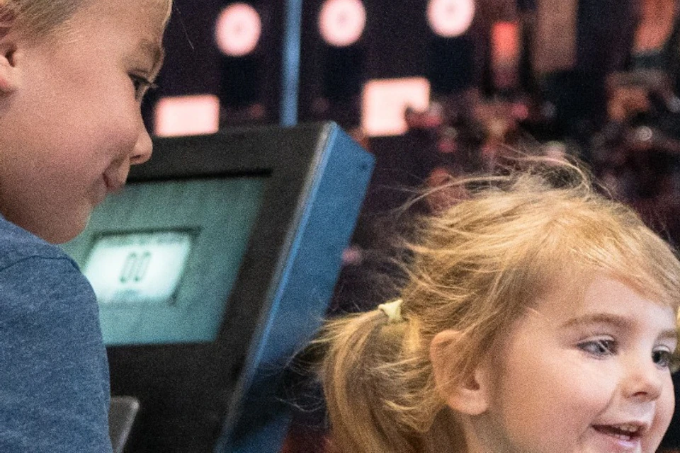 Two children interact with a large industrial wheel in a museum-like setting with machinery and a bright display in the background.