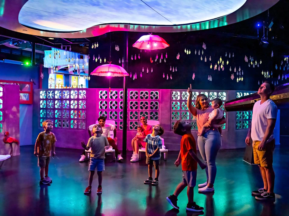 Children and adults look up in a brightly lit room with colorful lights, hanging pink umbrellas, and decorative wall panels.