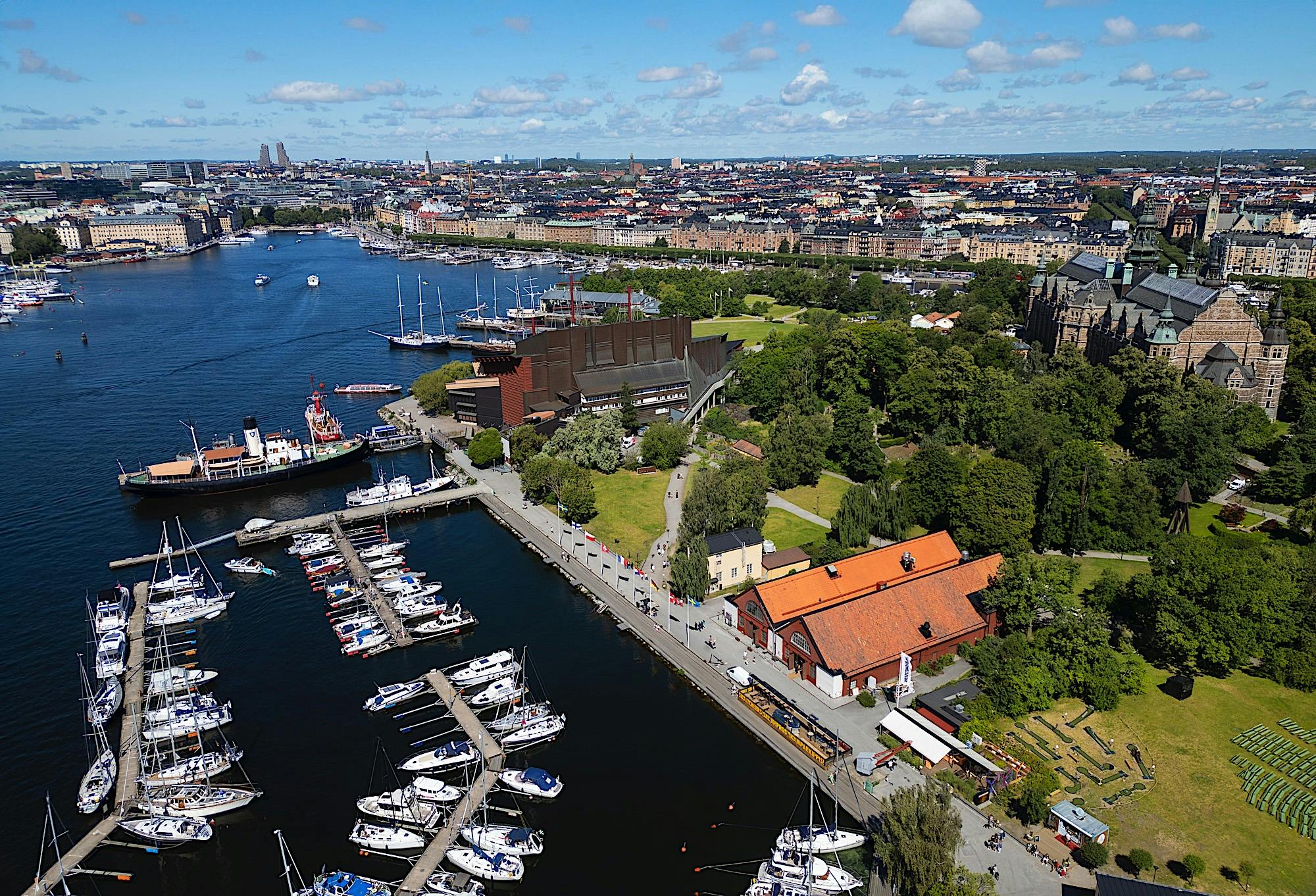 Vue aérienne d'un port de plaisance avec des bateaux, un parc et des bâtiments dans un paysage urbain sous un ciel bleu avec des nuages épars.