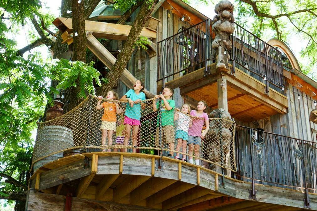 Children standing on a wooden balcony with netting, part of a multi-level wooden treehouse surrounded by greenery.
