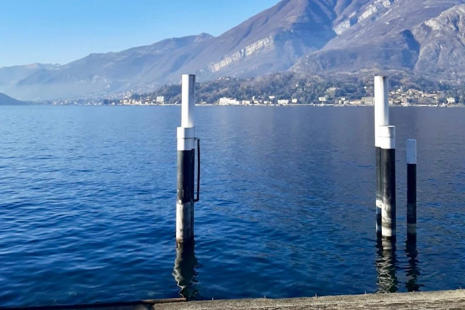 Two white posts stand in a calm lake with a village and mountains in the background under a clear blue sky.