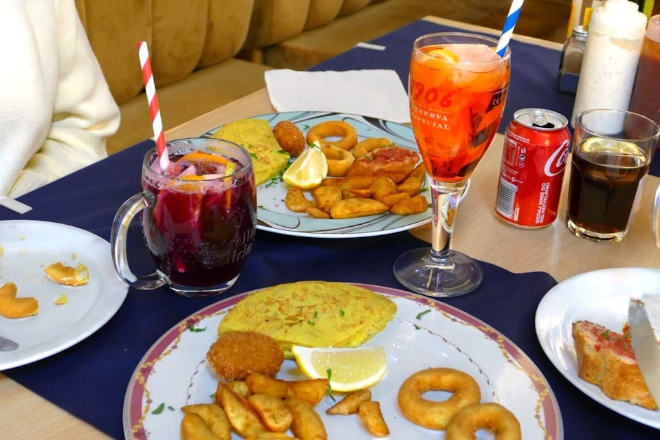 Two plates of assorted fried foods with lemon wedges, accompanied by colorful drinks and a can of soda on a table.