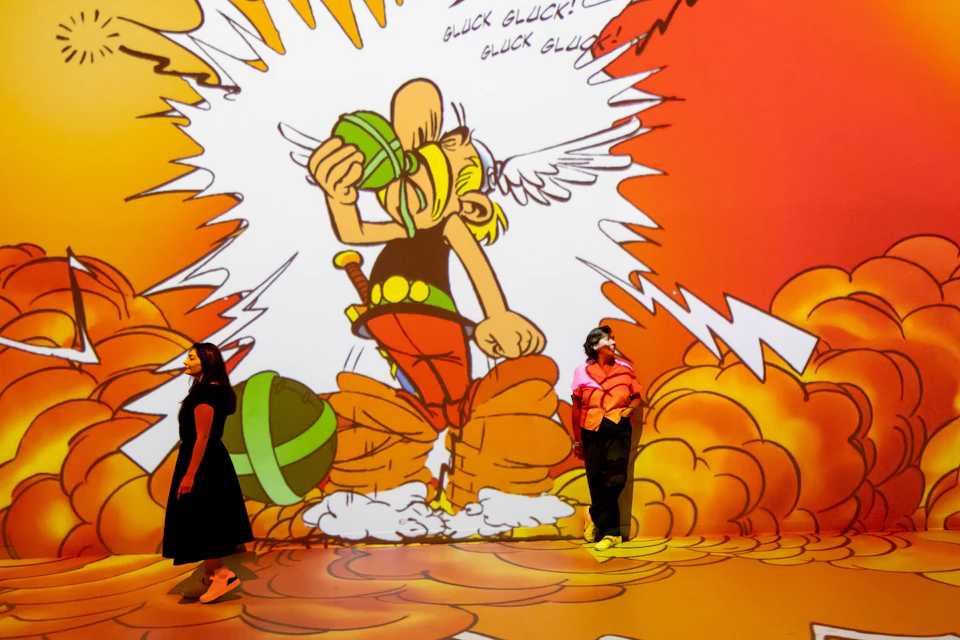 Two people walk in front of a large, colorful cartoon projection featuring a character drinking from a green container.