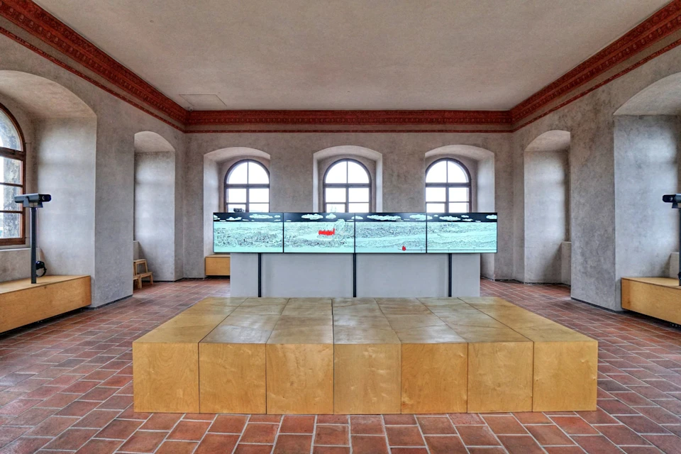 A room with tiled floor, three arched windows, a wall-mounted multi-screen display, and a wooden block structure in the foreground.