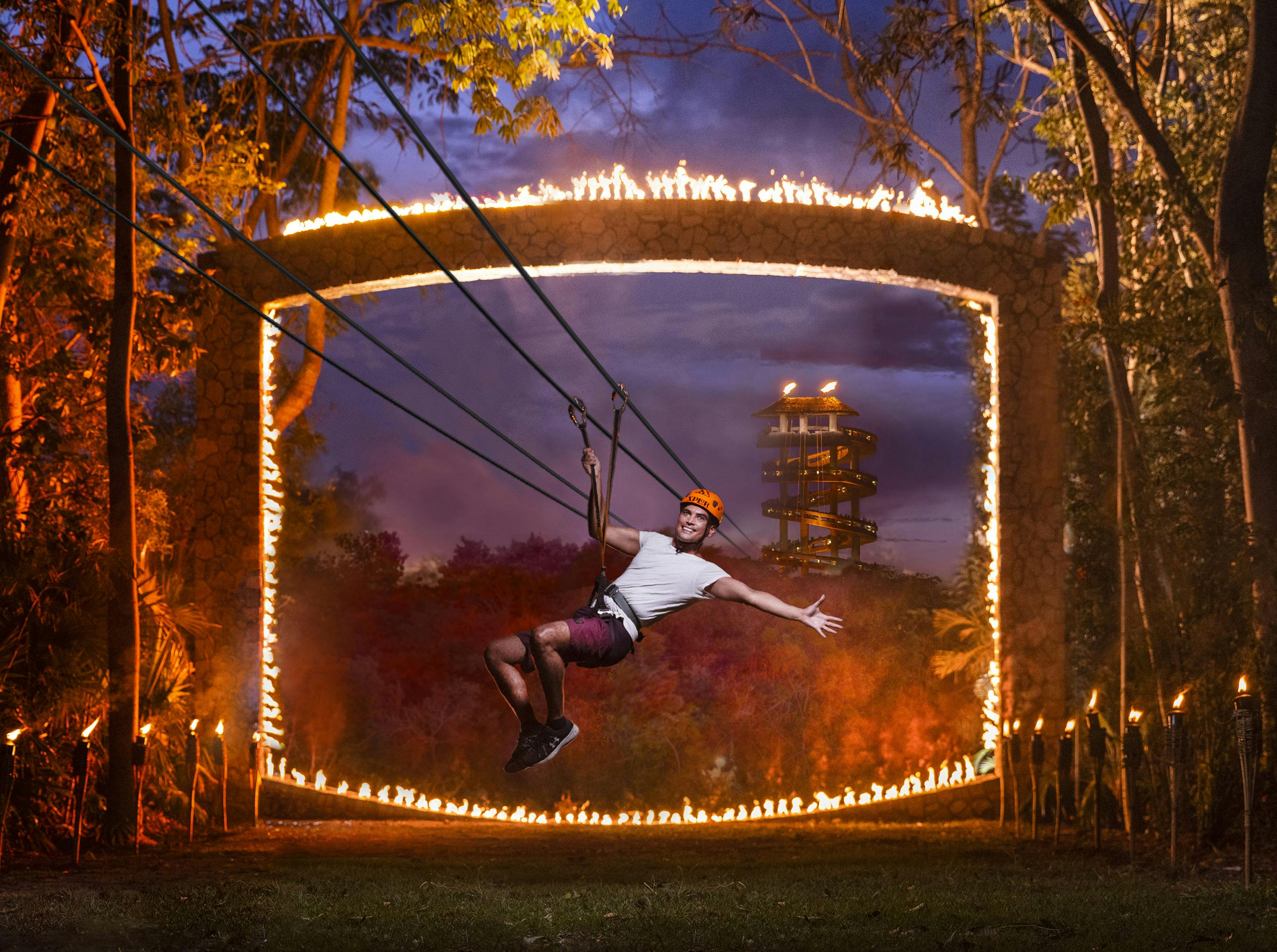 Une personne portant un casque fait de la tyrolienne dans un anneau de feu, avec une tour en spirale et des arbres en arrière-plan au crépuscule.