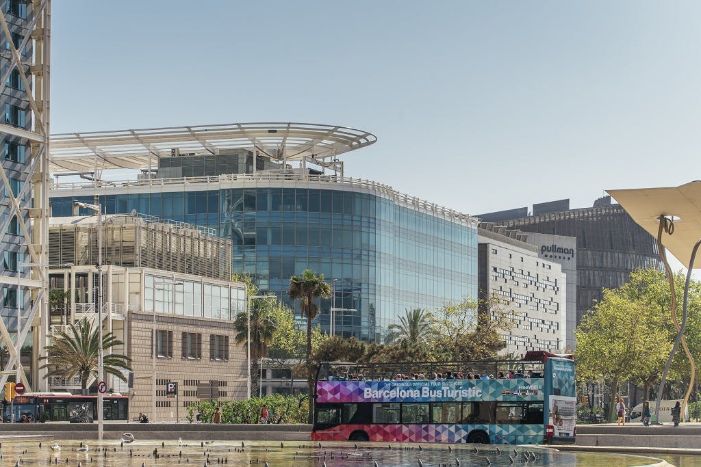 Un colorit autobús de dos pisos &#34;Barcelona Bus Turístic&#34; davant d&#39;edificis moderns de vidre i palmeres en un dia clar.