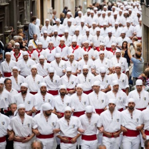 Un grande gruppo di persone in abiti bianchi coordinati con fusciacche e copricapi rossi che camminano in una strada stretta e affollata.