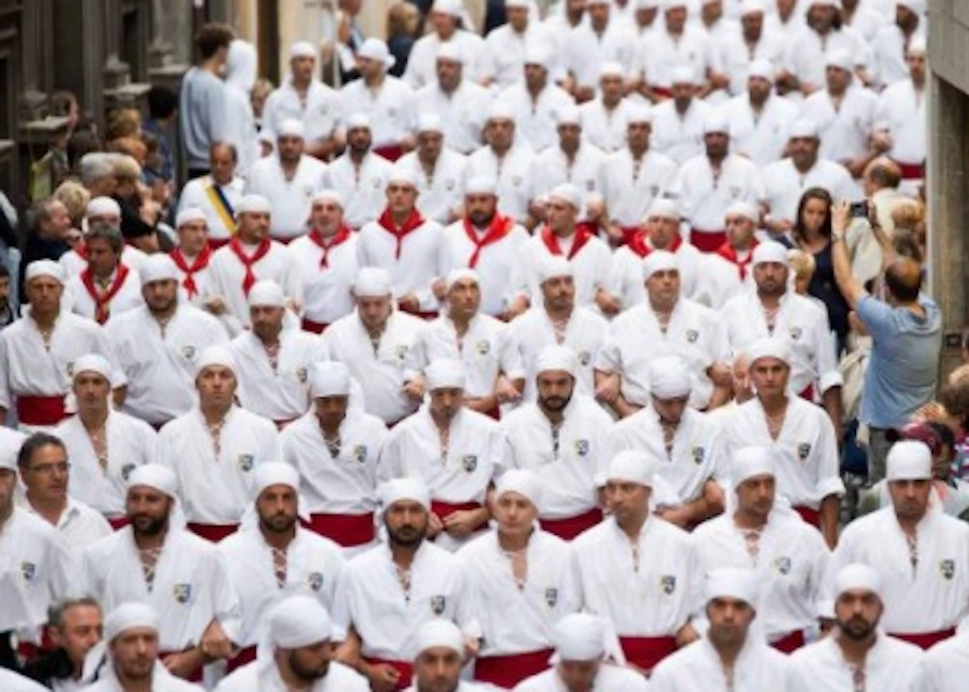 A large group of people in matching white outfits with red sashes and head coverings walking down a narrow, crowded street.
