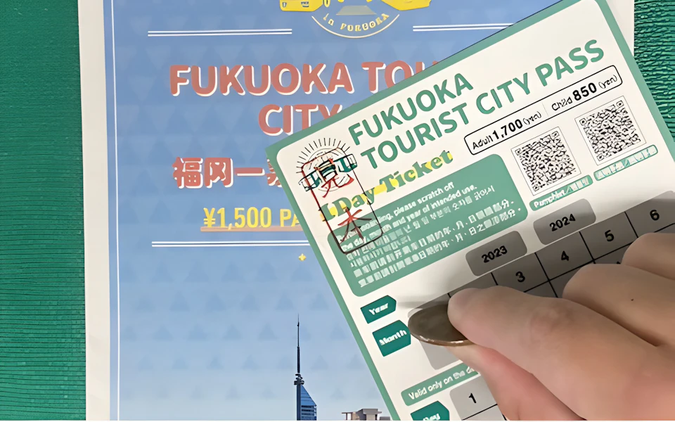 A hand scratching a Fukuoka Tourist City Pass ticket with a promotional poster in the background.