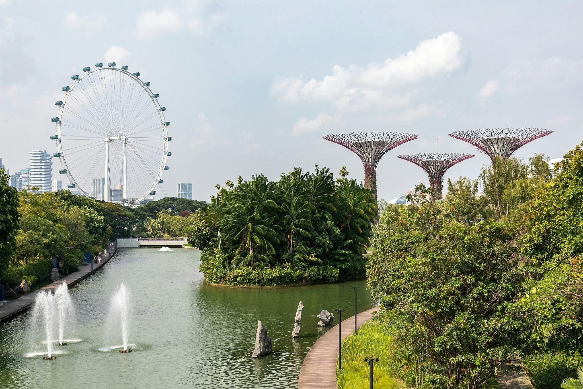 Large Ferris wheel and Supertrees amid lush greenery and fountains in a canal, with city buildings in the background.