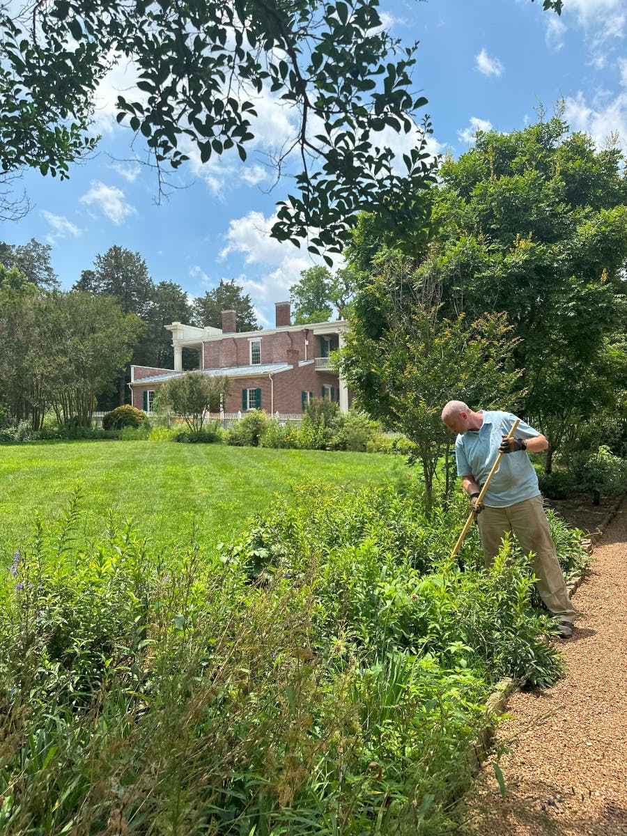 A person wearing a light blue shirt and gloves is gardening near a path. There's a large brick house and lush greenery in the background.