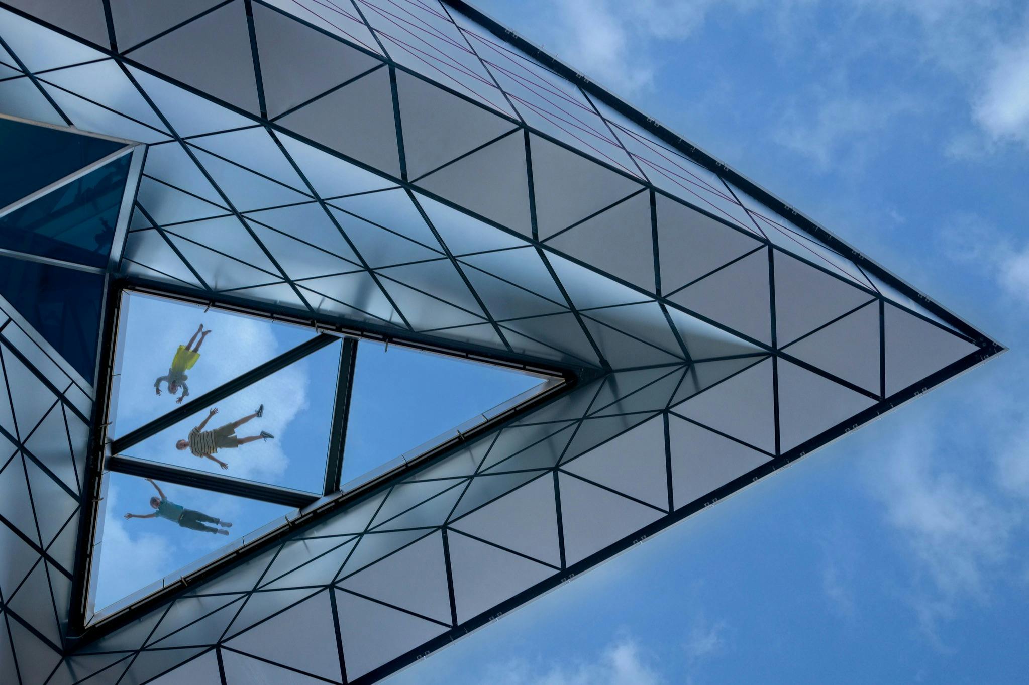 Geometrická skleněná konstrukce s trojúhelníkovými panely odrážejícími oblohu a zrcadlovým obrazem lidí vzhlížejících.