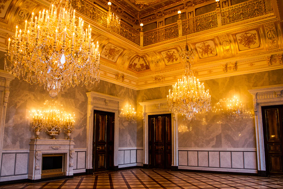 Elegant room with ornate chandeliers, golden ceiling decorations, wooden doors, and a checkered floor.