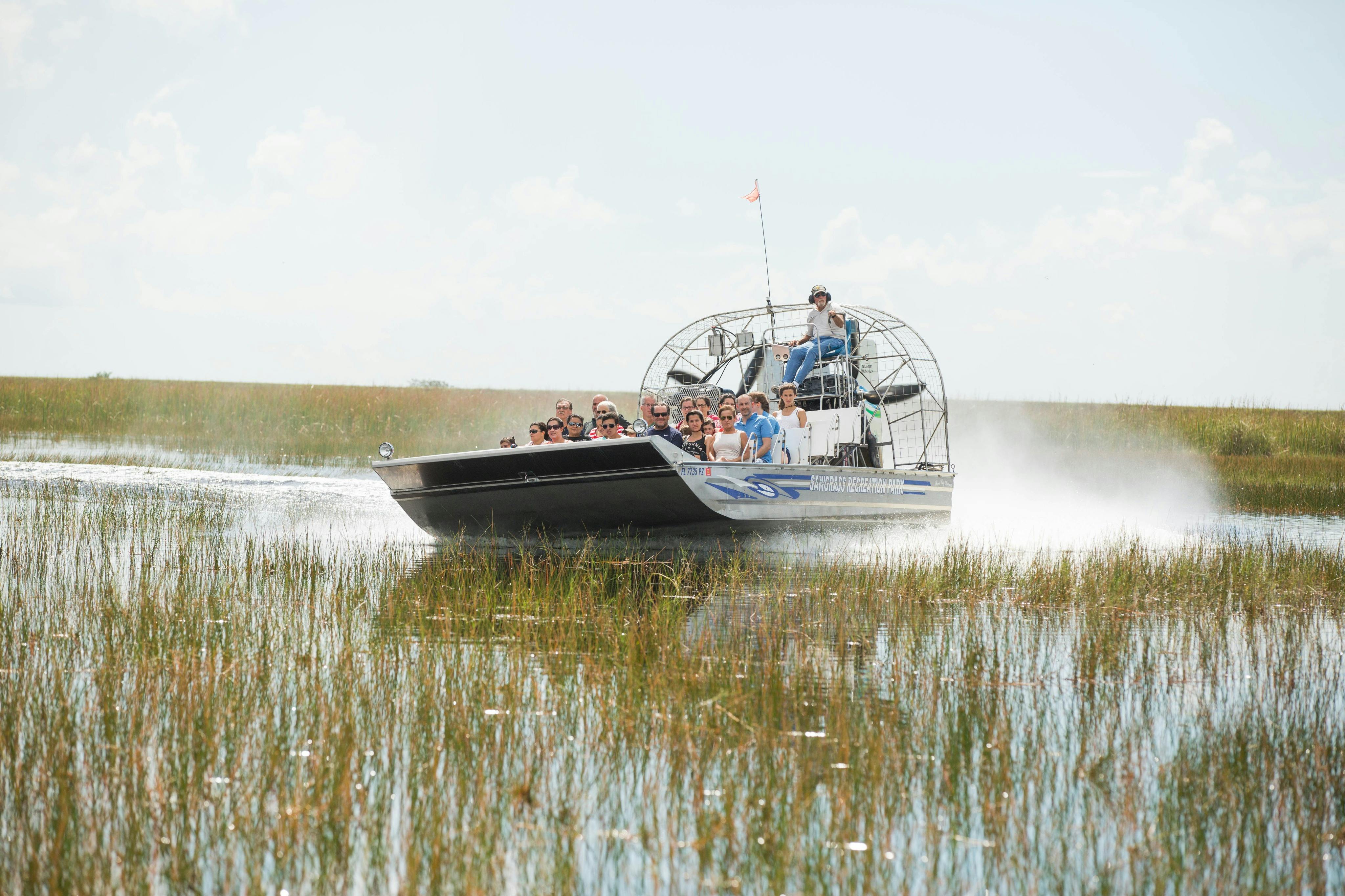 An airboat full of people glides through a marshy area under a clear sky, with a pilot steering at the back.