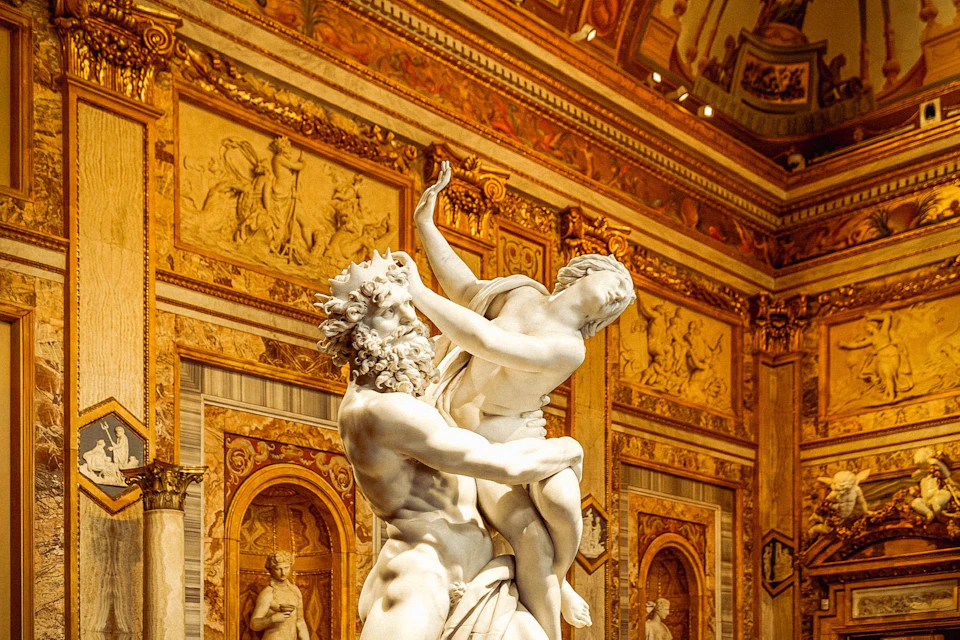 A marble statue of a bearded man holding a woman, set against an ornate, golden adorned wall with historical reliefs and columns.