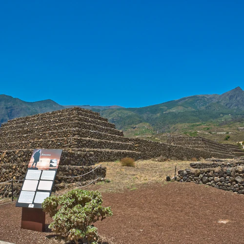 Steinpyramide mit umgebenden Mauern, Informationsschild im Vordergrund und entfernten Bergen unter einem klaren blauen Himmel.