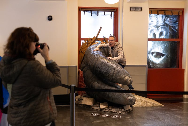 Ένας άντρας ποζάρει μέσα σε ένα μεγάλο έκθεμα σχεδιασμένο να μοιάζει με το χέρι του Κινγκ Κονγκ που τον αρπάζει, με φόντο το πρόσωπο του Κινγκ Κονγκ.