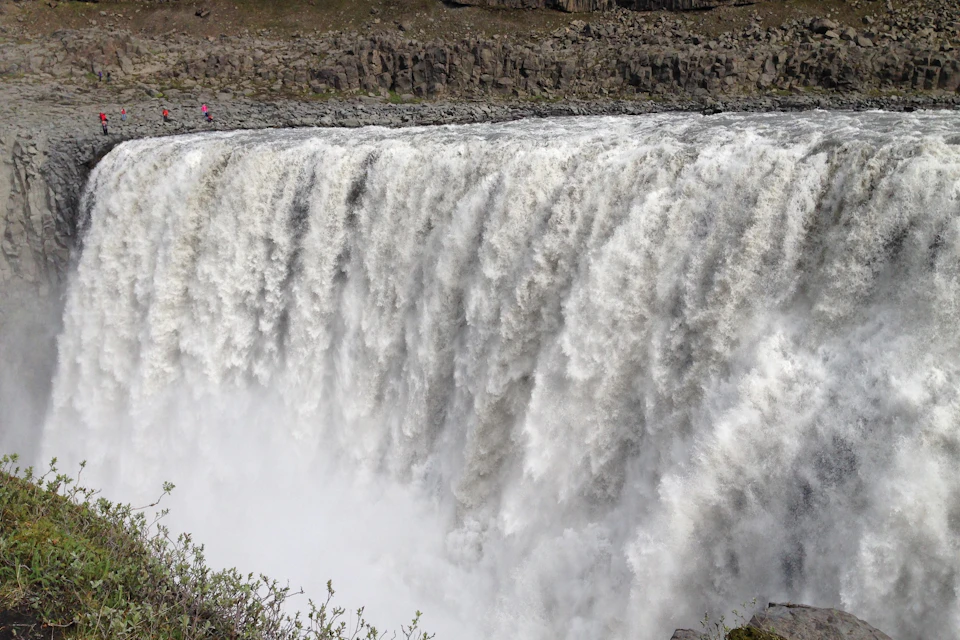 Massive waterfall cascading over a rocky cliff with mist rising at the base; two small figures on the edge for scale.